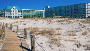 A beachside hotel with a wooden boardwalk leading to the entrance, a large building with multiple balconies, and a smaller building with a blue roof.