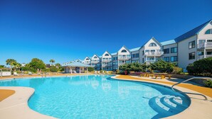 A large swimming pool surrounded by multi-story buildings with balconies, a covered area with lounge chairs, and a clear blue sky.