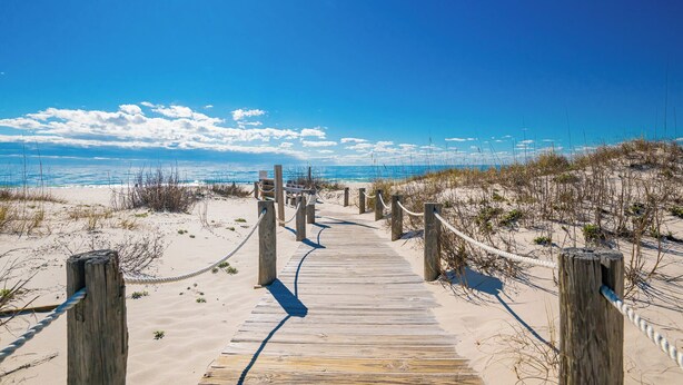 A wooden boardwalk leading to a beach with clear skies and distant ocean views.