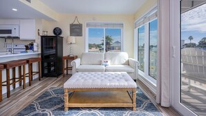 A compact living space with a kitchenette, a white sofa, a tufted ottoman, and large windows offering a view of palm trees and buildings.