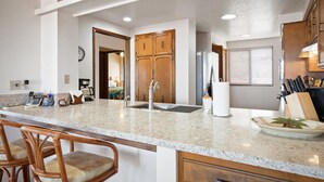 A kitchen with a granite countertop, wooden cabinets, a sink, and a breakfast bar with chairs. - Aloha Condos, Kona Coast Resort, Condo 1-304, Ocean View (Kailua Kona)
