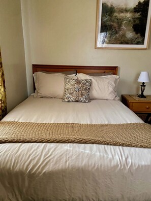 Una cama bien hecha con una almohada estampada, una manta tejida y una lámpara de noche sobre una mesita de madera.