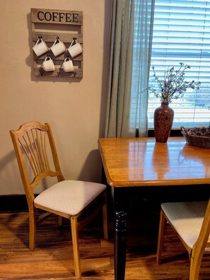 Une table à manger en bois avec des chaises, un panneau indiquant un café, un vase avec des fleurs et une fenêtre avec des stores.