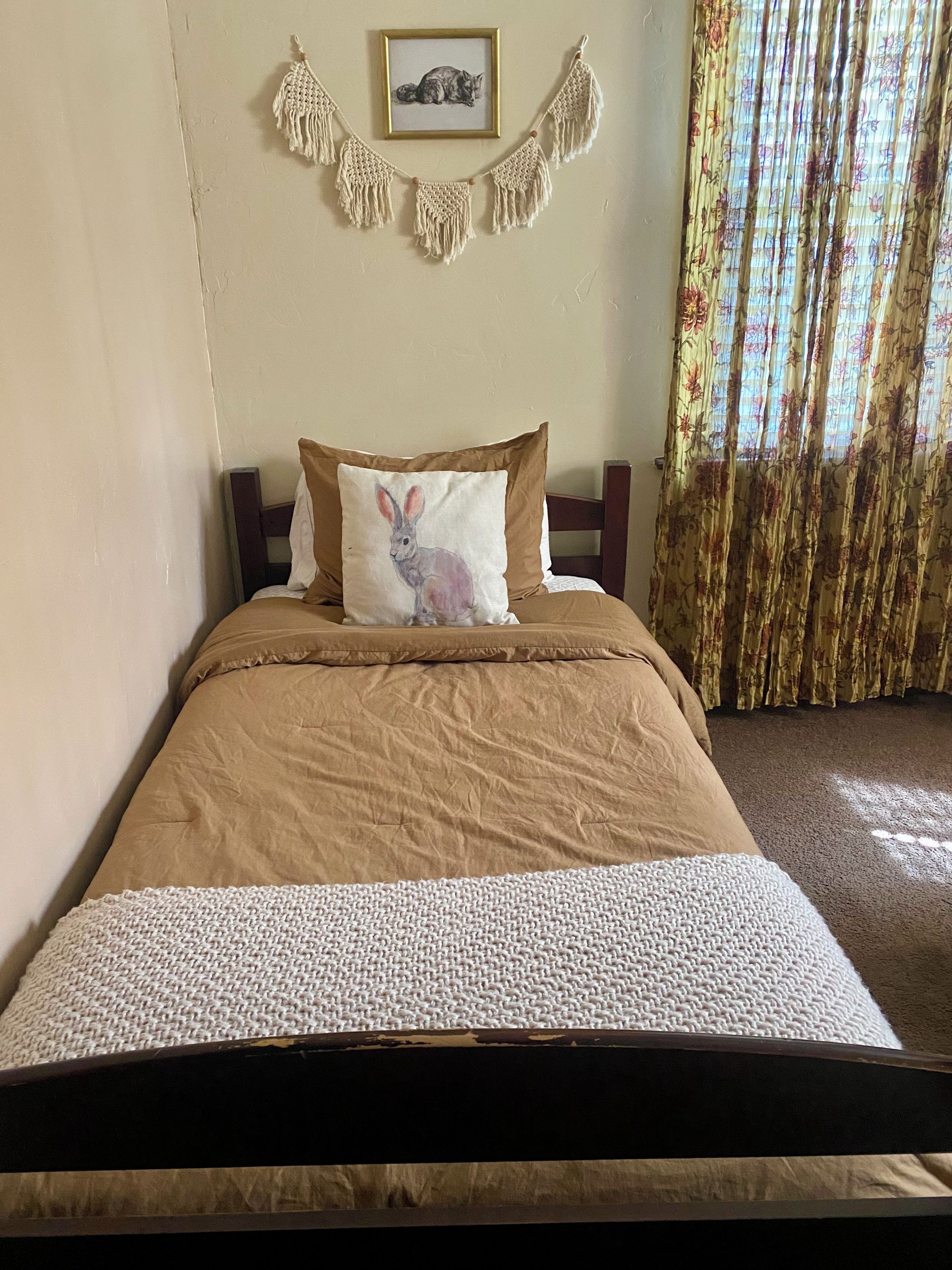 A bed with a brown bedspread, a white pillow with a rabbit design, and a macrame wall hanging above.