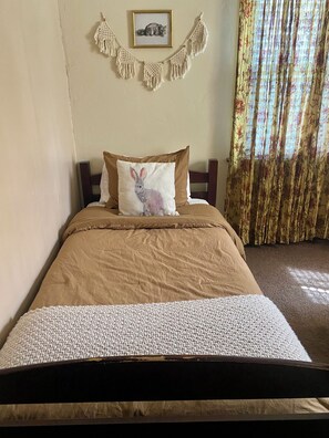 Una cama con una colcha marrón, un almohadón blanco con diseño de conejo y un tapiz de macramé en la pared.