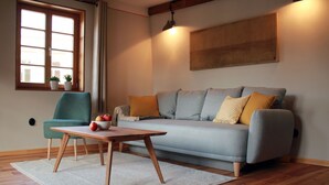 A living room with a sofa, a table with fruit, a chair, a window with plants, and a painting on the wall.