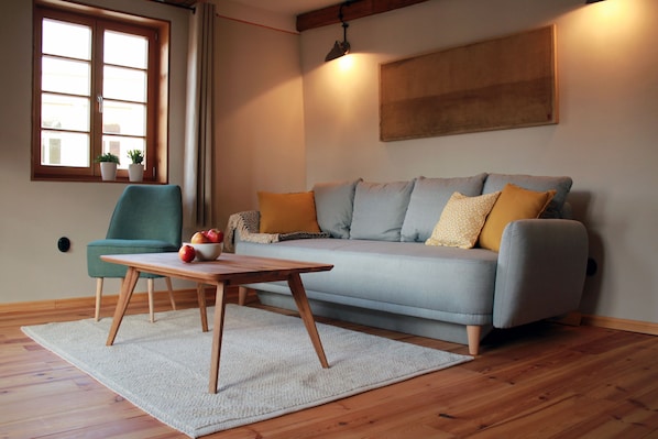 A living room with a sofa, a table with fruit, a chair, a window with plants, and a painting on the wall.