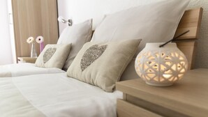 Una cama bien hecha con edredón blanco y dos almohadas con diseño de corazones. Una lámpara blanca con motivos geométricos está sobre la mesita de noche.