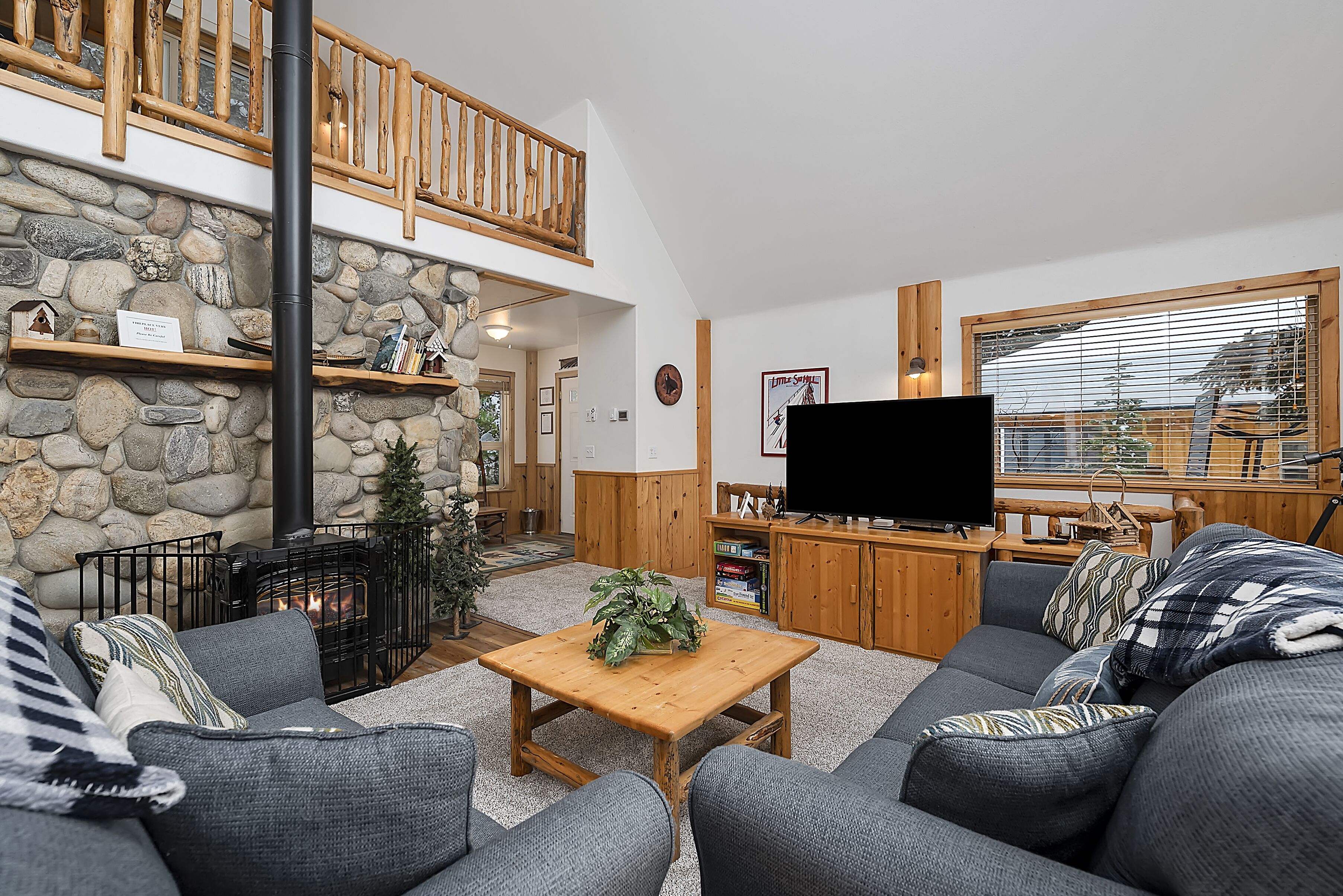 A cozy living room with a stone fireplace, a wooden TV stand, and a large window with blinds.