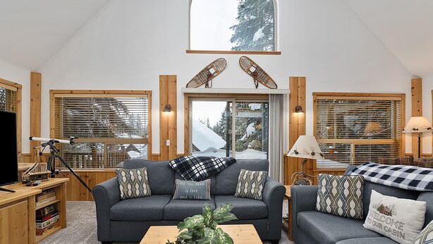 A cozy living room with a dark grey sofa, wooden accents, and a large window with blinds.