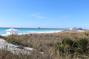Une plage avec une petite construction blanche, une jetée qui s’avance dans l’eau et une zone herbeuse au premier plan.