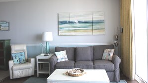 Een woonkamer met een grijze bank, een witte salontafel, een klein bijzettafeltje en een schilderij van zeilboten aan de muur.
