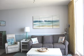 Uma sala de estar com um sofá cinzento, uma mesa de centro branca, uma mesinha lateral e um quadro com barcos à vela na parede.