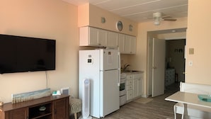 Une cuisine compacte avec des meubles blancs, un four encastré et un réfrigérateur. On y trouve une table à manger avec un saladier, un meuble en bois et un ventilateur sur pied.
