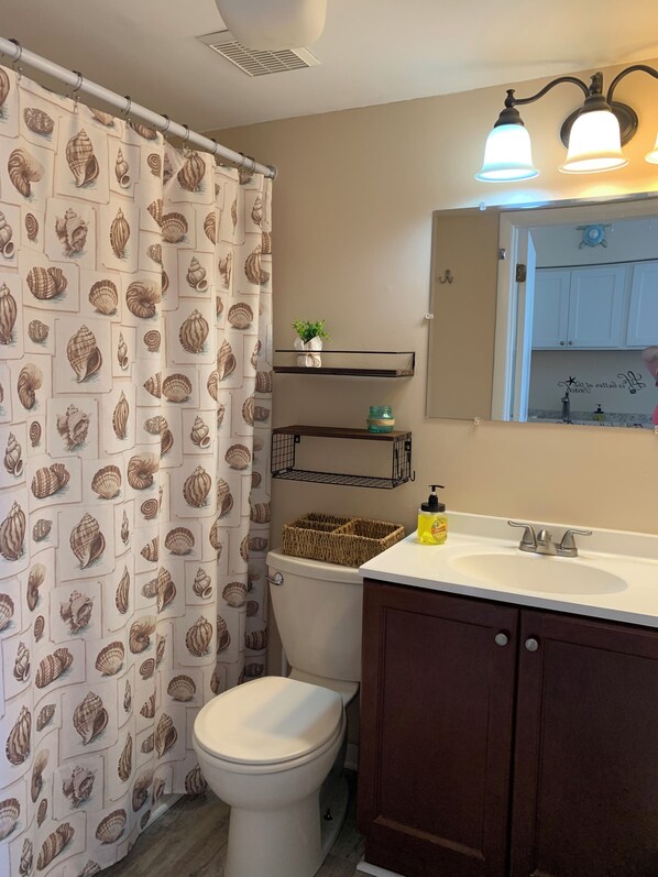 A bathroom with a toilet, a sink with a mirror above it, a shower curtain with a shell pattern, and a shelf with a plant and a basket. - Comfy condo 1 block to beach and boards. Perfect for your family beach vacation! (Wildwood)