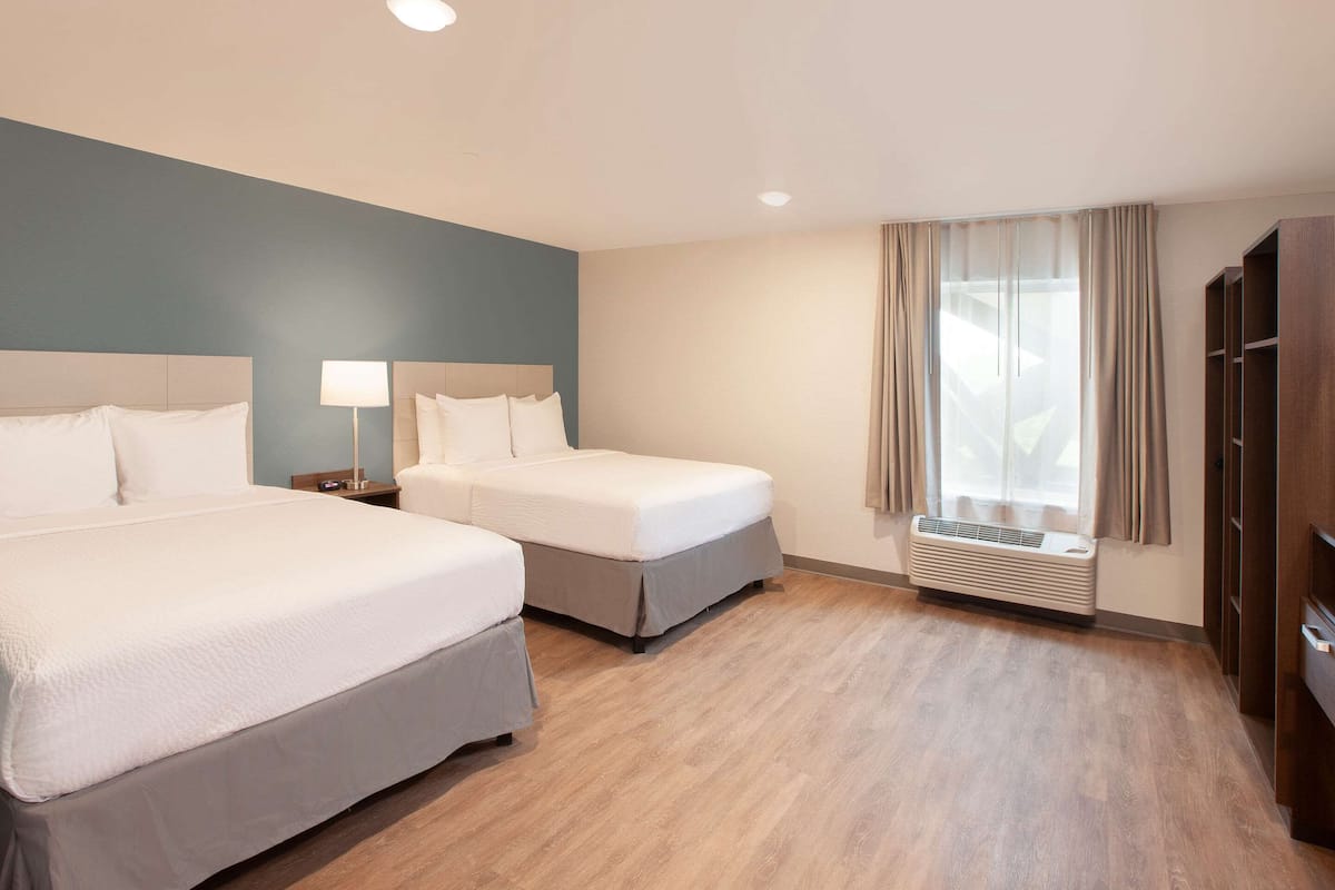 Ein Hotelzimmer mit zwei Betten, einem Nachttisch mit Lampe, einem Fenster mit Vorhängen und einer fest installierten Klimaanlage.