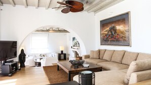 Una sala de estar moderna con un sofá grande, una mesita de centro y un televisor. Hay un cuadro en la pared y un ventilador de techo.