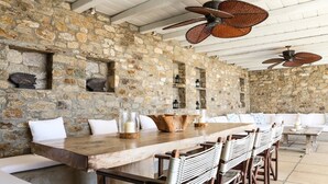 Un comedor con paredes de piedra, una larga mesa de madera, sillas tejidas y ventiladores de techo.