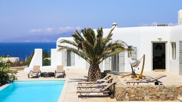 Zona junto a la piscina con vistas al mar, una palmera y un edificio blanco con puertas y ventanas.