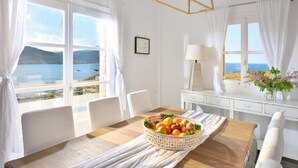 A room with a wooden dining table, white chairs, a fruit basket, and a view of the sea through large windows. - Villa Juliet (Mykonos)