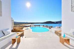 Zona de piscina con vista al mar y colinas, suelo de piedra y mobiliario blanco.