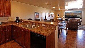 Cocina con encimeras de granito, lavamanos de acero inoxidable y lavavajillas. Hay un comedor con mesa y sillas, y una sala de estar con sofá y vistas al exterior.