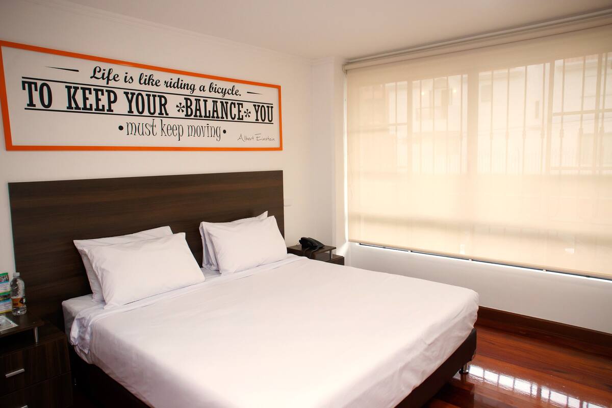 A neatly made bed with white linens in a hotel room, a wooden headboard, a bedside table with a phone, and a large window with blinds.