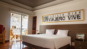 Une chambre spacieuse avec un grand lit, un plancher en bois, un balcon avec vue et une inscription sur le mur qui dit : « UN TOURISTE REGARDE, UN VOYAGEUR VIT ».