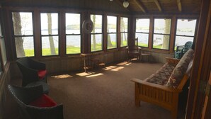 Una cabaña con ventanas amplias que dan a un lago, amueblada con un banco y sillas de madera.