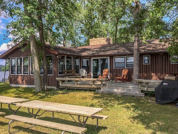A rustic cabin with a deck, wooden benches, and Adirondack chairs.
