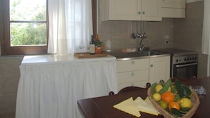 A small kitchen with white cabinets, a sink, and a stove. There is a window with curtains, a table with a fruit basket, and a tray with wine glasses and a flower. - La Quercia-Erica - One Bedroom Apartment, Sleeps 5 (Val Canina)