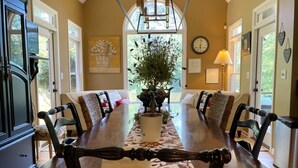 A dining room with a long table, chairs, a chandelier, a potted plant, and a clock on the wall.