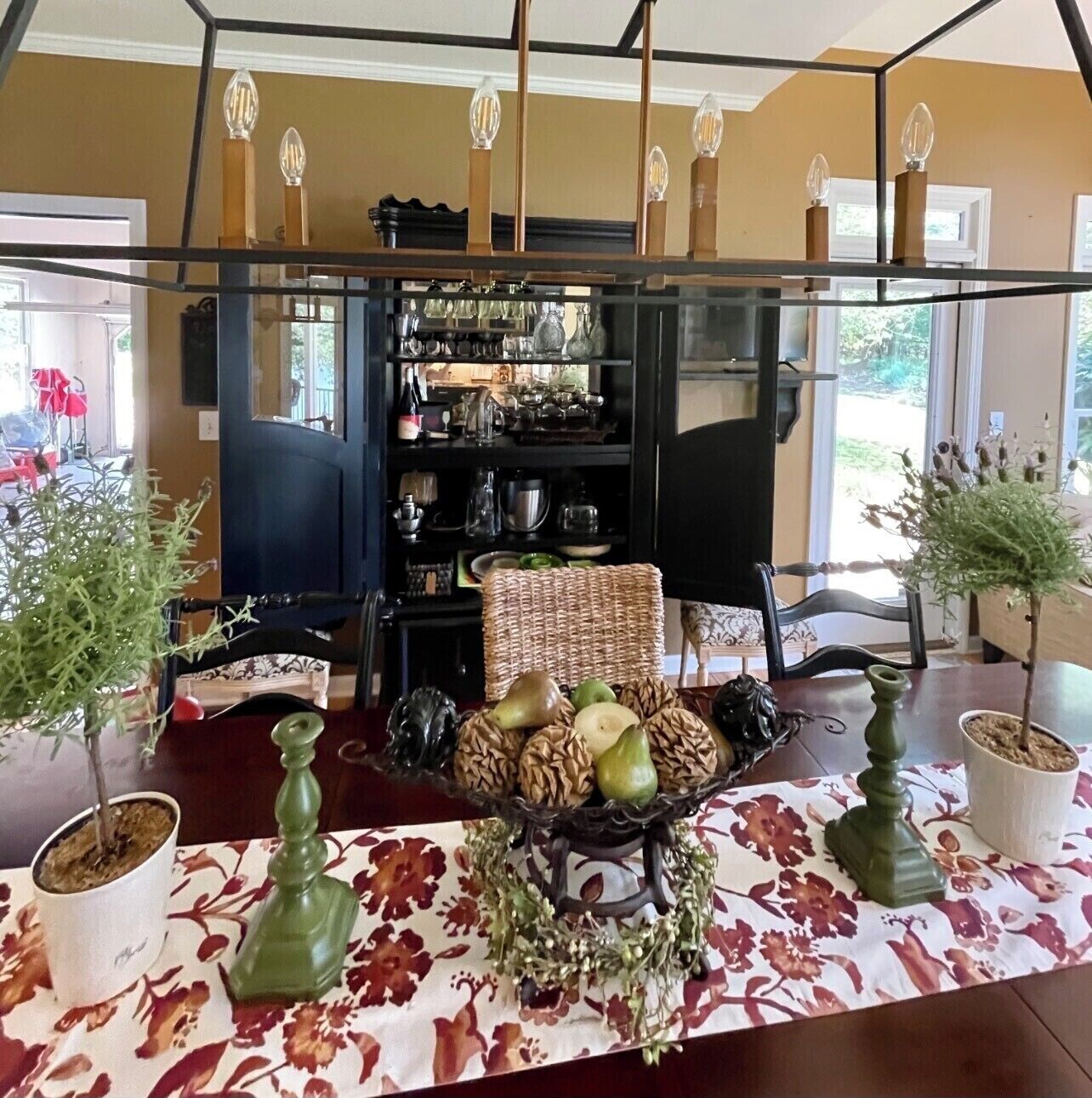 Un comedor con una mesa lista para dos personas, con mantel floreado, un centro de mesa con frutas y frutos secos, y dos macetas.