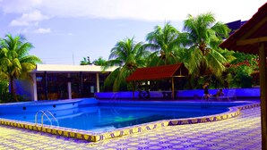 Une piscine dotée d’une terrasse carrelée, entourée de palmiers et bordée d’un bâtiment au toit rouge.
