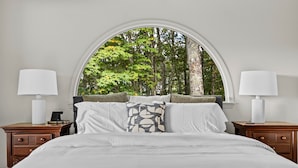 Un dormitorio con un amplio ventanal arqueado, una cama con sábanas blancas, dos mesitas de noche con lámparas y vistas a árboles.