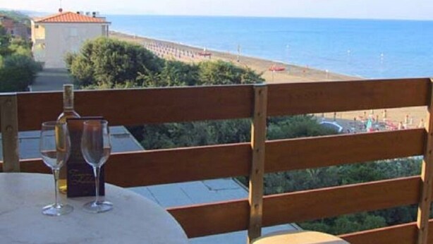 Una terrazza con tavolo, due bicchieri e una bottiglia, vista sulla spiaggia e sul mare.