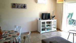 A living room with a TV on a white cabinet, a dining table with chairs, and a large window with a view of greenery. - House with private pool, not shared between Vaison la Romaine and Nyons (Saint-Maurice-sur-Eygues.)