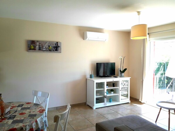 Una sala de estar con una televisión sobre un mueble blanco, una mesa de comedor con sillas y un ventanal con vistas a la vegetación.