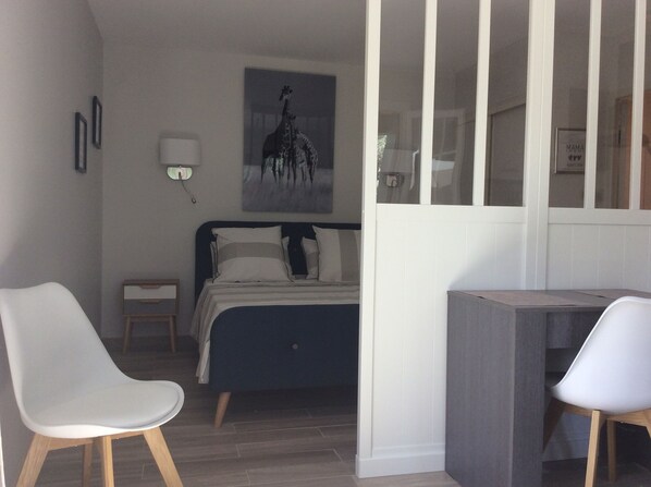 A bedroom with a bed, two chairs, a desk, a nightstand, a wall-mounted lamp, and a framed picture.