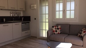 A modern kitchen with white cabinets, a built-in oven, and a black countertop. There is a grey sofa with a polka dot cushion and a small table in front. - Air-conditioned studio with charming little garden in Villeneuve les Avignon (Villeneuve-Lès-Avignon)