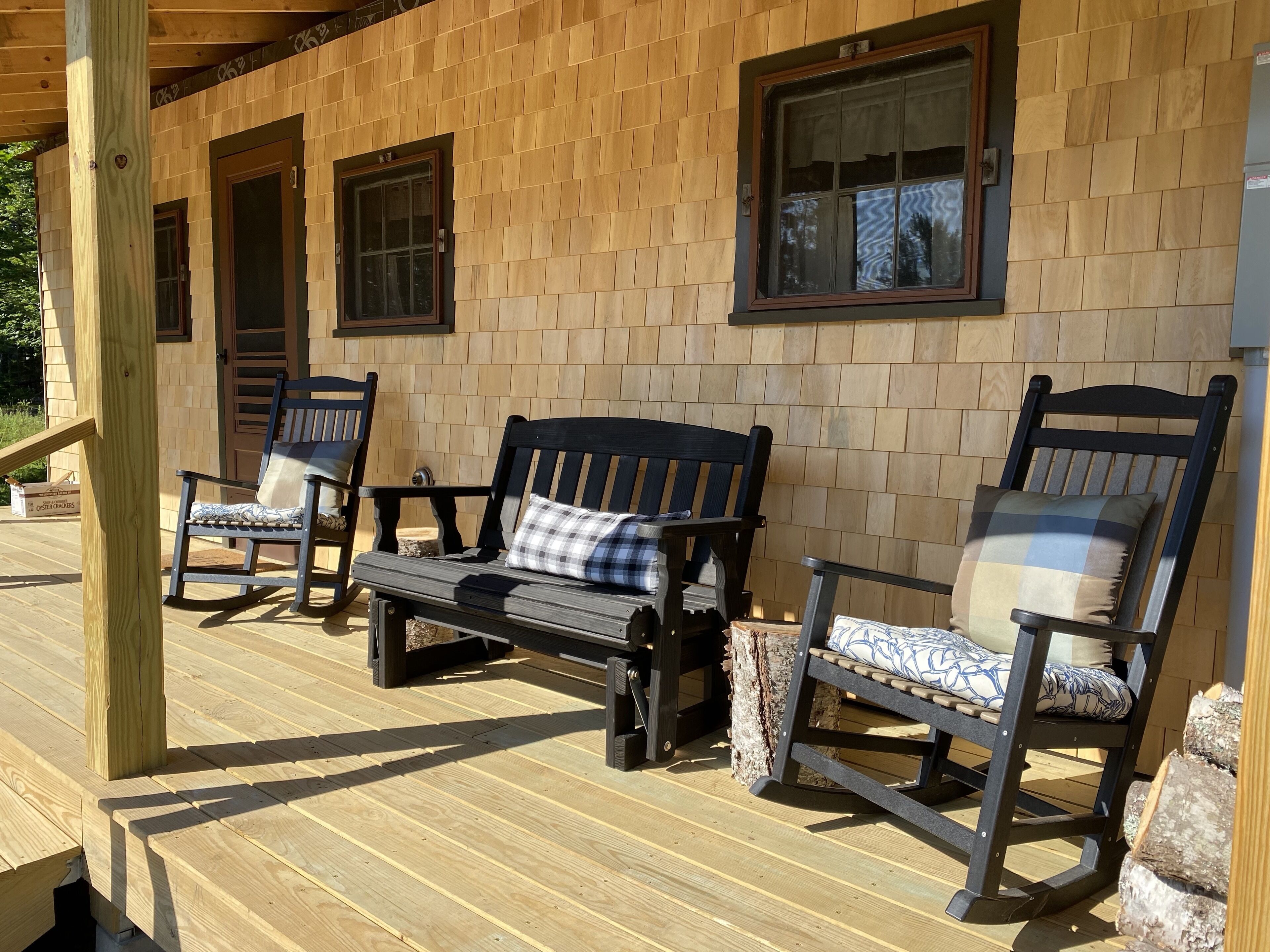 Une terrasse en bois avec un banc et deux fauteuils à bascule, une porte en bois et une fenêtre.