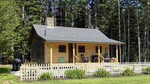 A log cabin with a porch, a white picket fence, and a small garden area. - A Historic Storybook Maine Cottage (New Harbor)