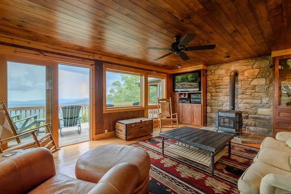 Cozy cabin interior with a rustic charm, featuring a fireplace, leather sofas, and a large window offering a view of the surrounding landscape.
