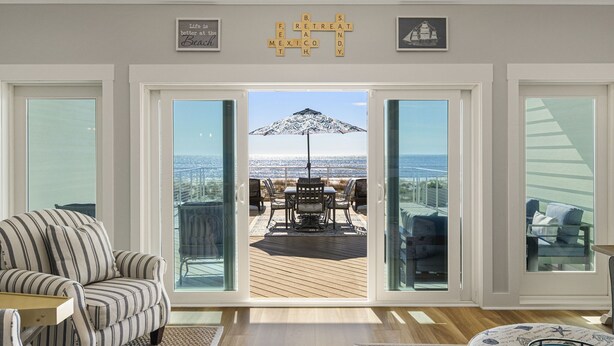 Un salón amplio con un gran ventanal corredera que da a un patio exterior con mesa y sillas, y vistas al océano.