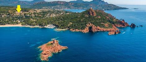 An aerial view of a coastal area with rocky terrain, clear blue waters, and a small island with a tower.