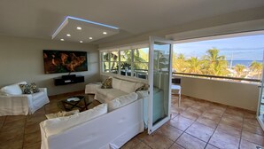 A spacious living room with a white sofa, a flat-screen TV mounted on the wall, and a large window offering a view of palm trees and the ocean. - BEACHFRONT > 5 STAR > 2 LEVEL PENTHOUSE VILLA > MAGNIFICENT PANORAMIC VIEWS  (Barrio Las Cabezas, Fajardo)