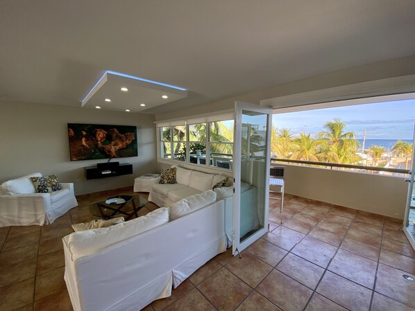 Un amplio salón con un sofá blanco, un televisor de pantalla plana montado en la pared y un ventanal con vistas a palmeras y al océano.