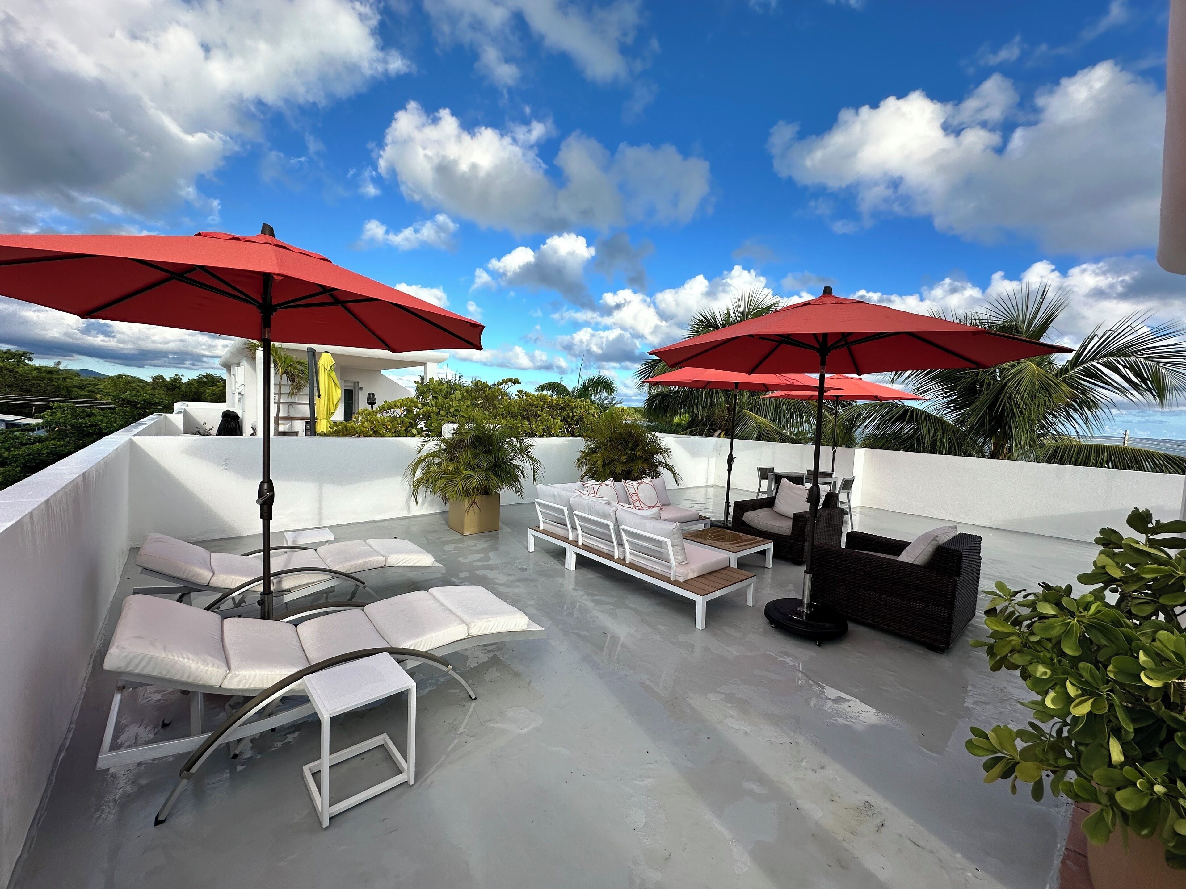 A rooftop patio with white furniture, red umbrellas, and a view of the ocean.