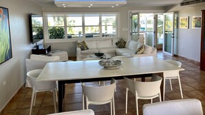 A modern living room with a white sofa, a dining table, and a TV. There are large windows with a view of palm trees outside. - BEACHFRONT > 5 STAR > 2 LEVEL PENTHOUSE VILLA > MAGNIFICENT PANORAMIC VIEWS  (Barrio Las Cabezas, Fajardo)