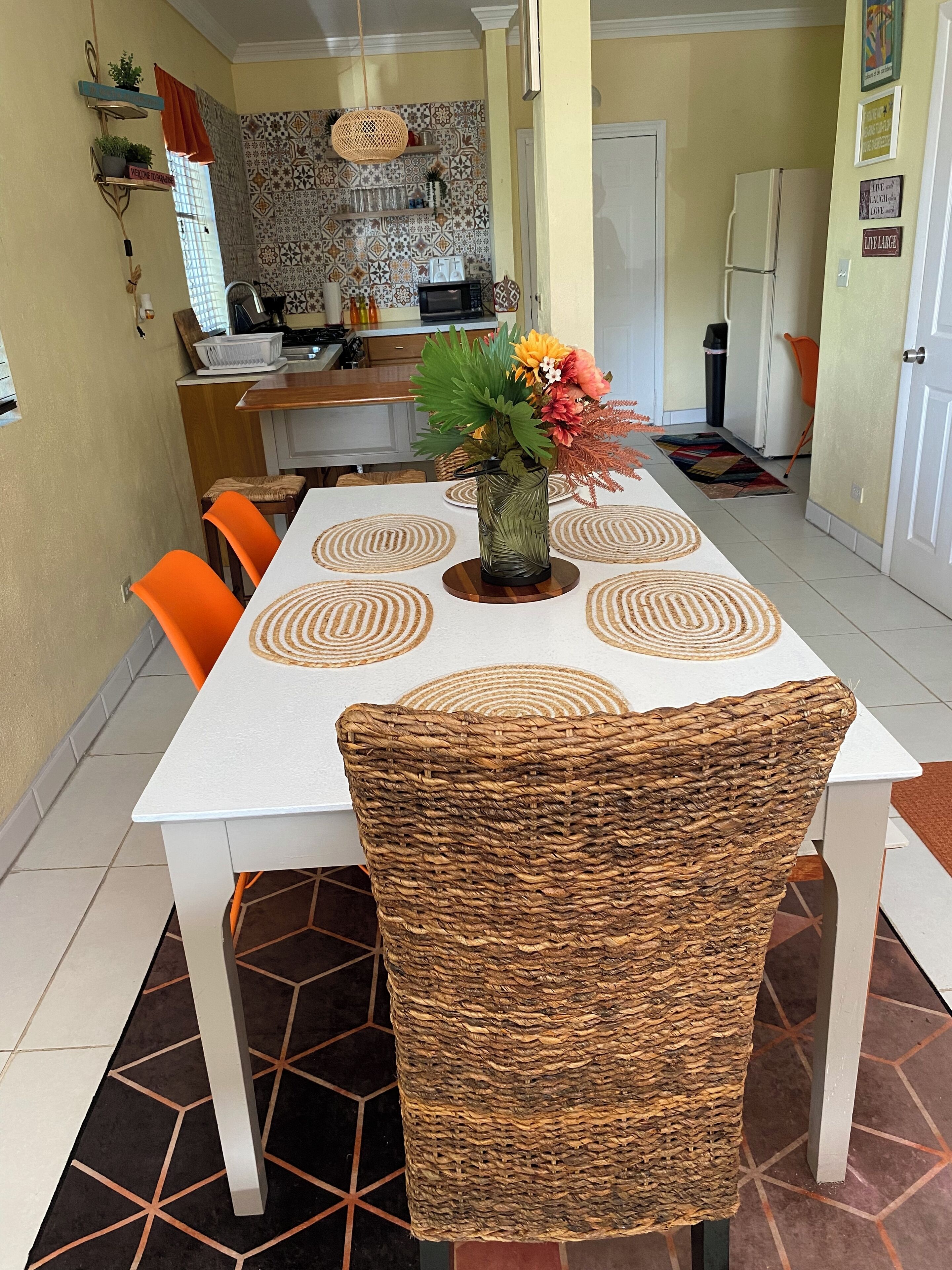 A dining area with a white table, orange chairs, and a woven chair. A vase with flowers is on the table. The kitchen is visible in the background with patterned tiles.